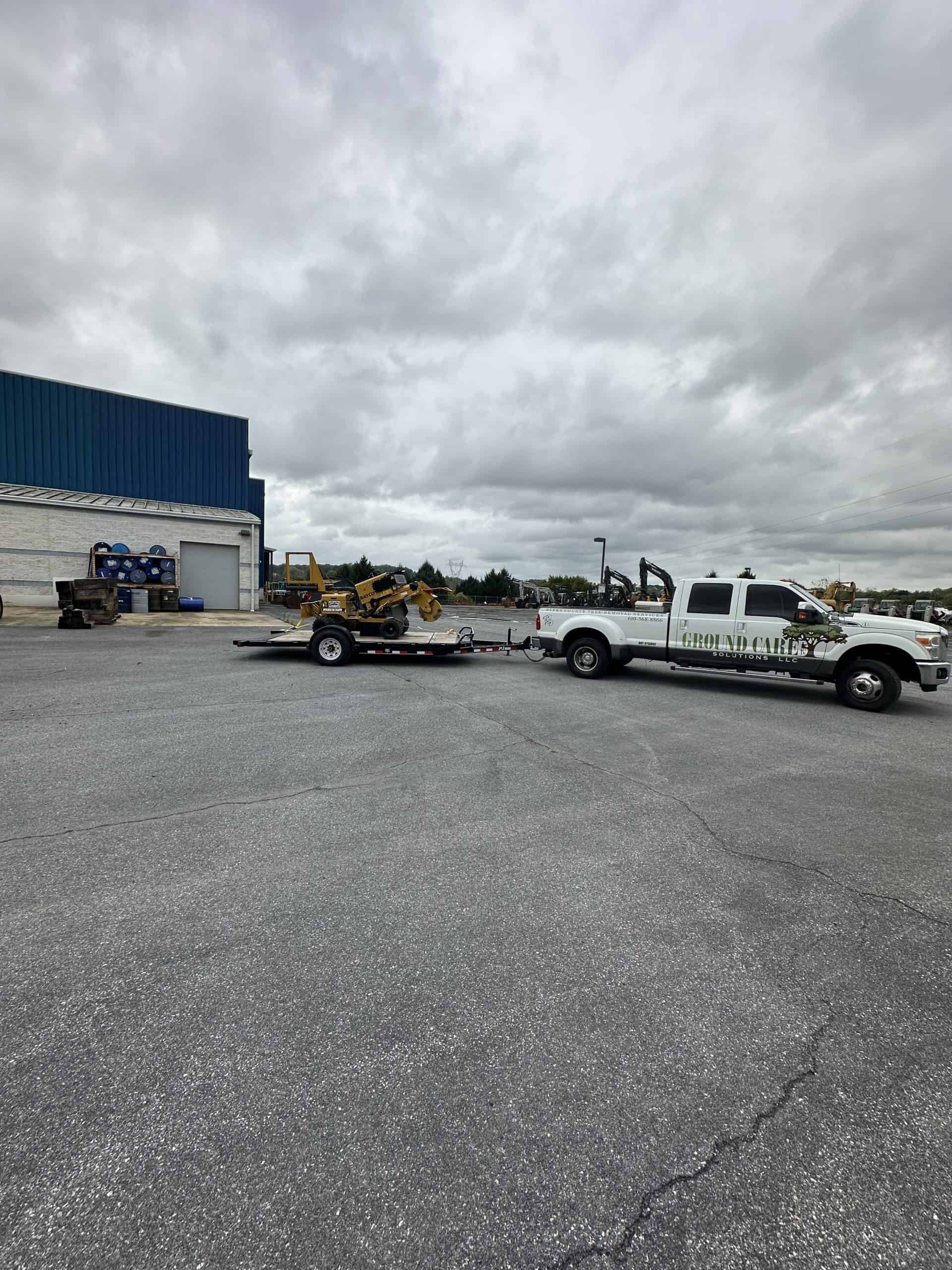 A white pickup truck with "Ground Care" written on the side is towing a flatbed trailer with a yellow construction vehicle on it. The scene is set in a parking lot with a cloudy sky overhead.