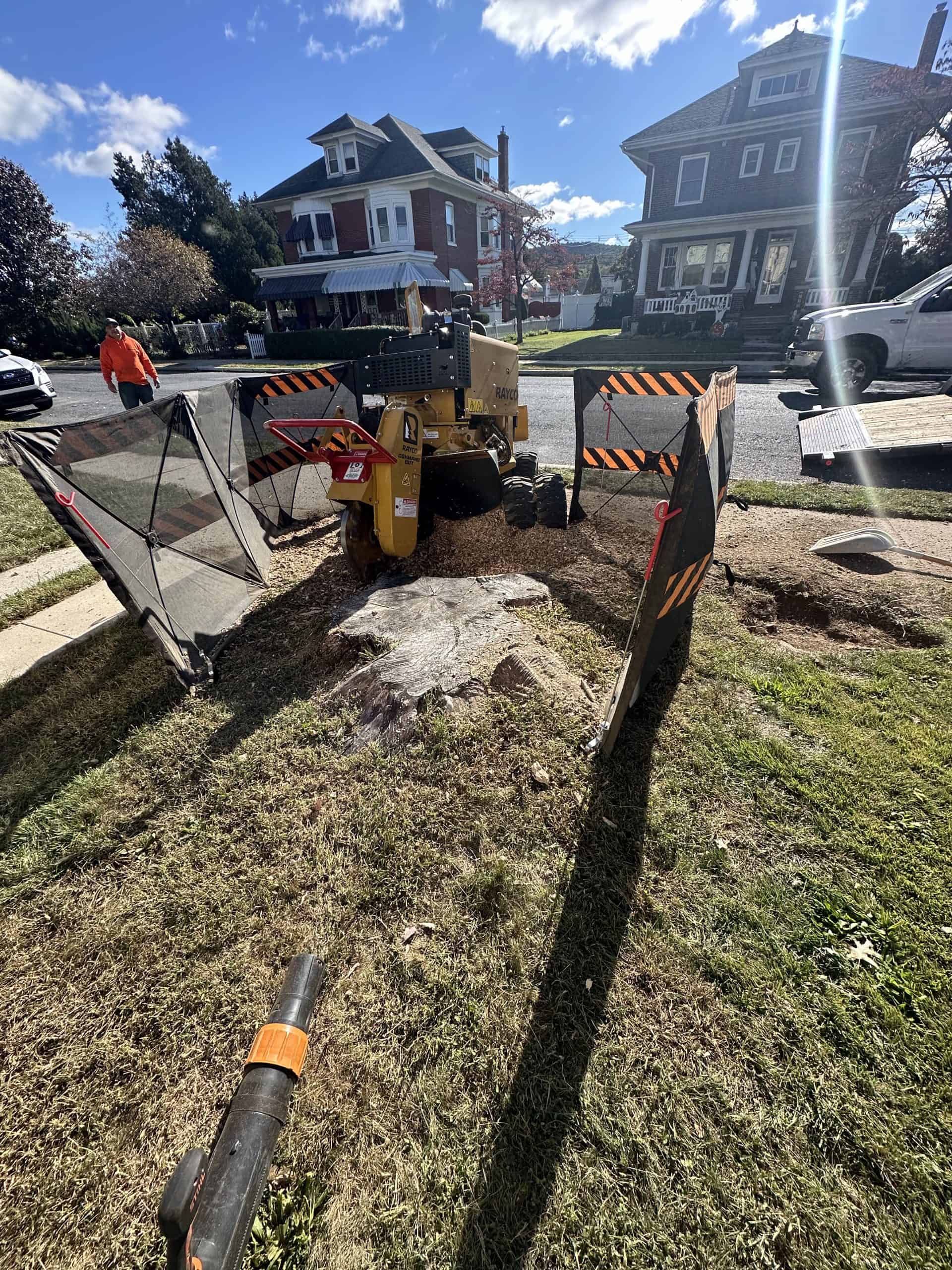 A stump grinder is positioned over a tree stump on a grassy lawn, surrounded by safety barriers. In the background, there are residential houses, a person wearing an orange shirt, and a parked white truck under a blue sky with fluffy clouds.
